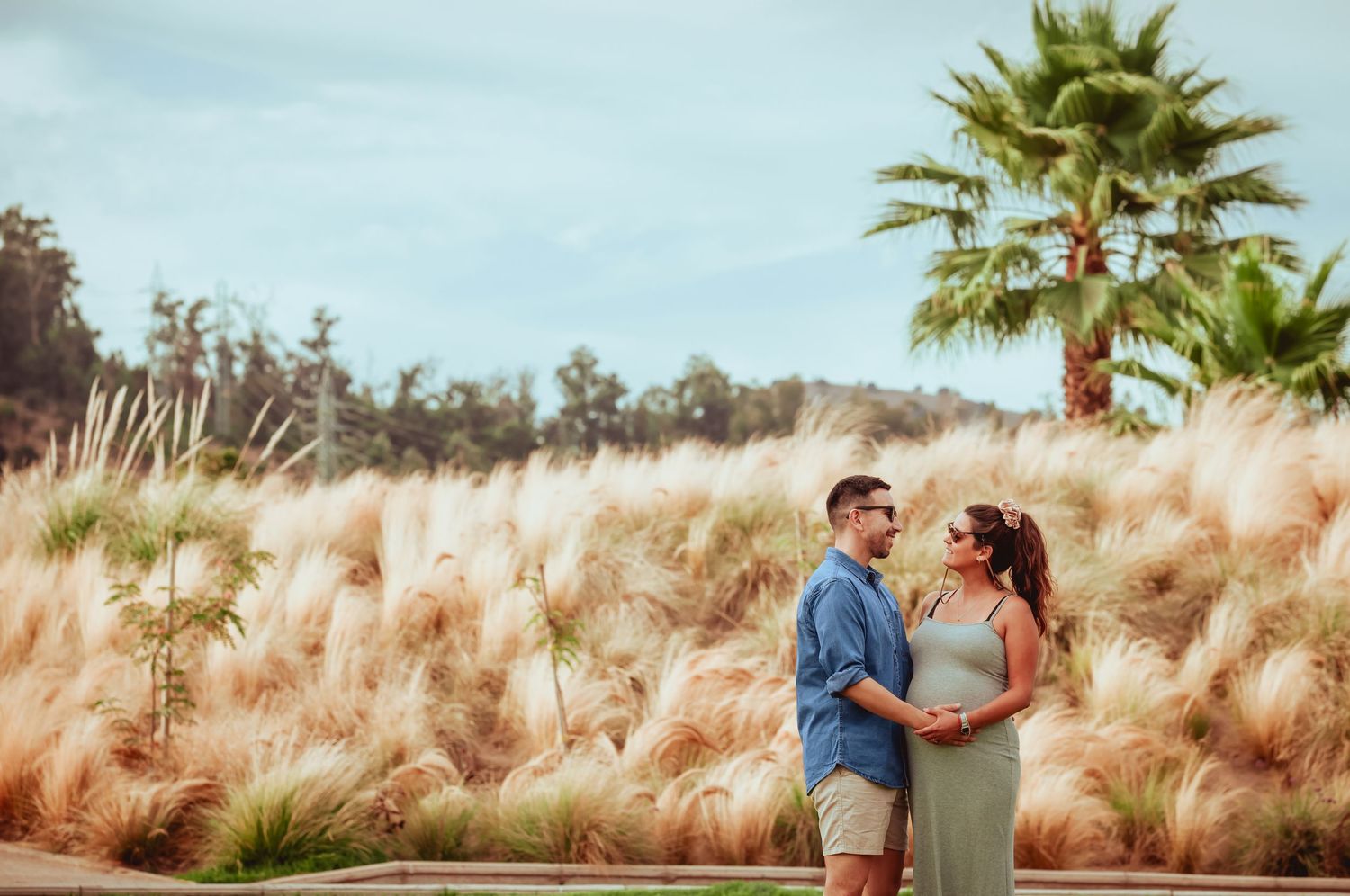 Expecting couple holding each other tenderly on green lawn with pampas grass and palm tree backdrop.