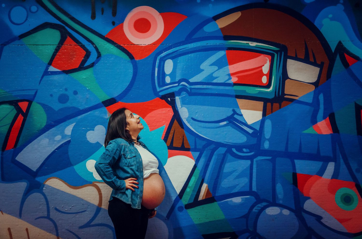 Pregnant woman in denim jacket gazing upward against a vivid blue graffiti mural backdrop.