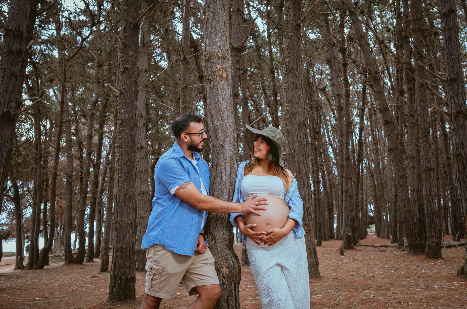 Couple touching pregnant belly together in pine tree forest setting.