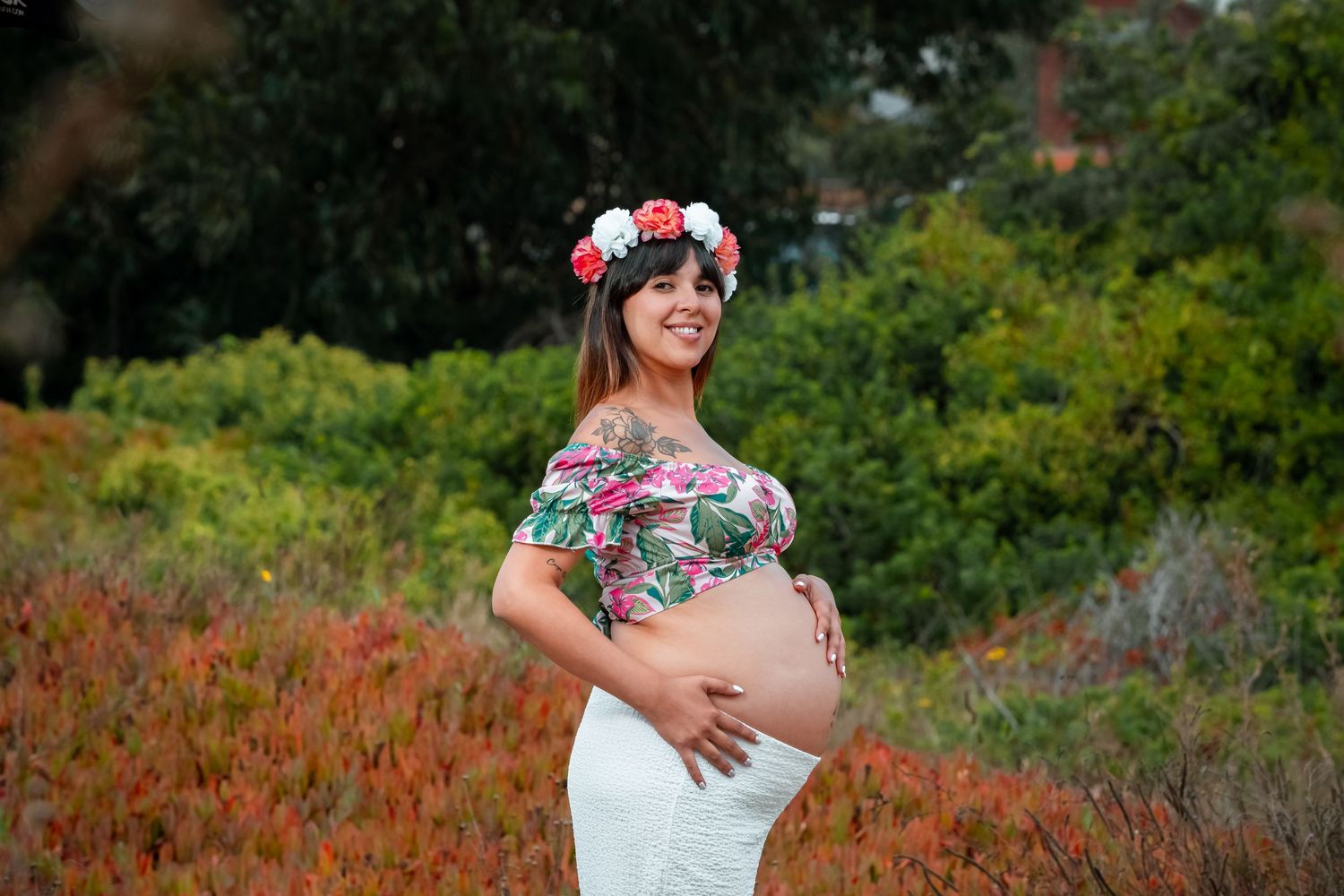 Smiling pregnant woman in floral crop top and white skirt with flower crown outdoors.