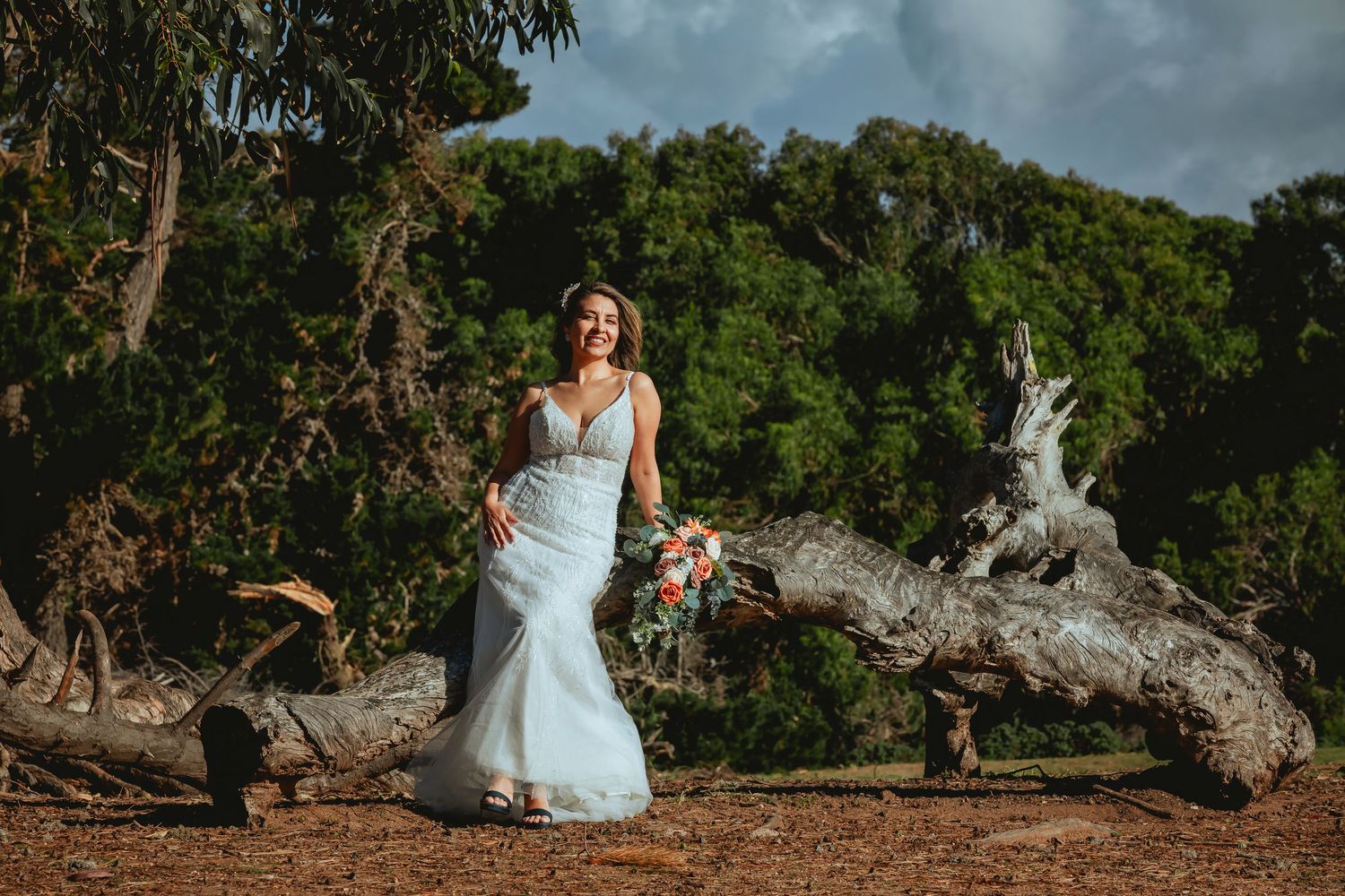 Smiling bride in a flowing light blue wedding gown holding a bouquet while posing on a weathered driftwood log with lush green trees behind.