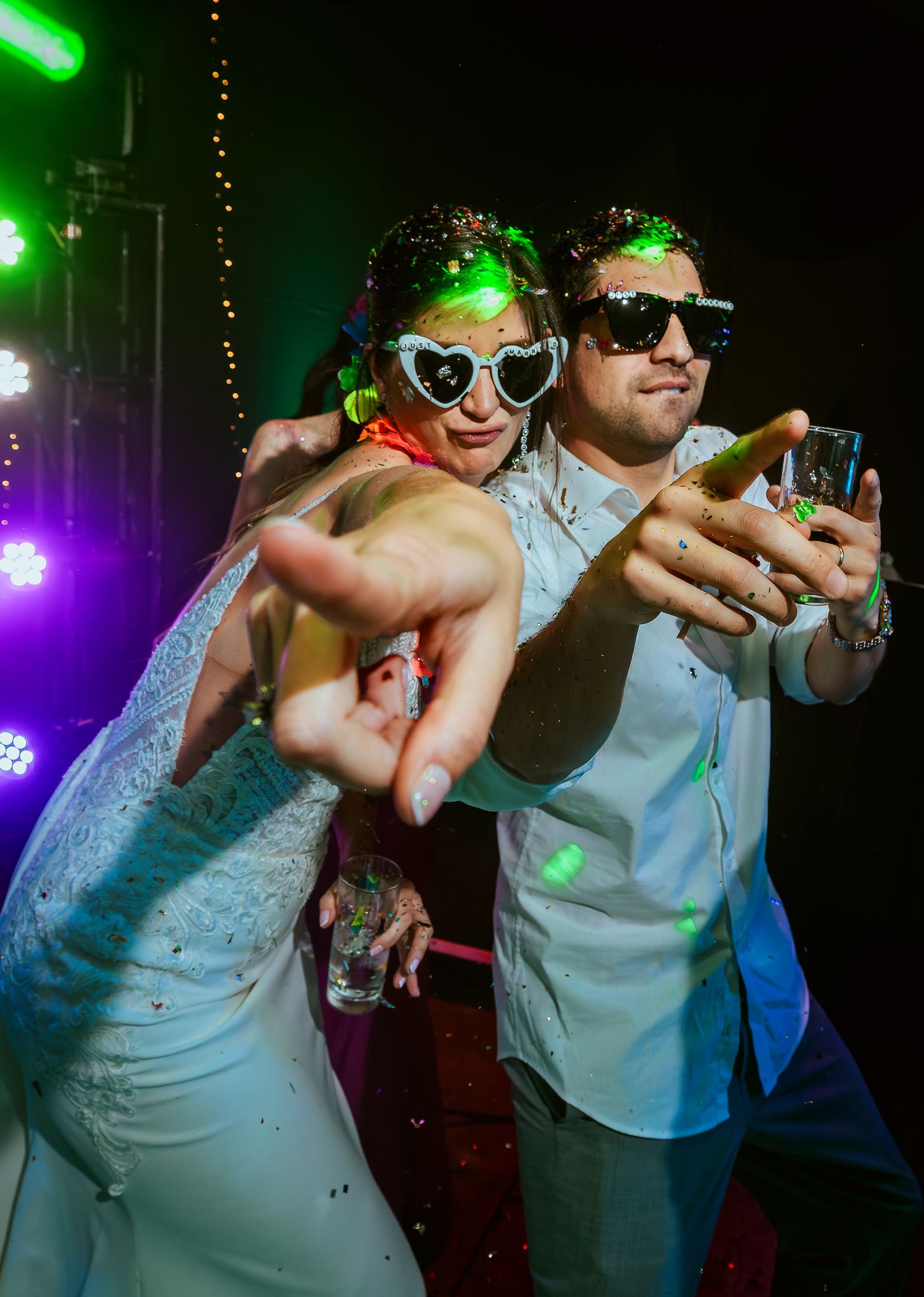 Couple in sunglasses striking fun poses at a wedding reception party with colorful stage lights.