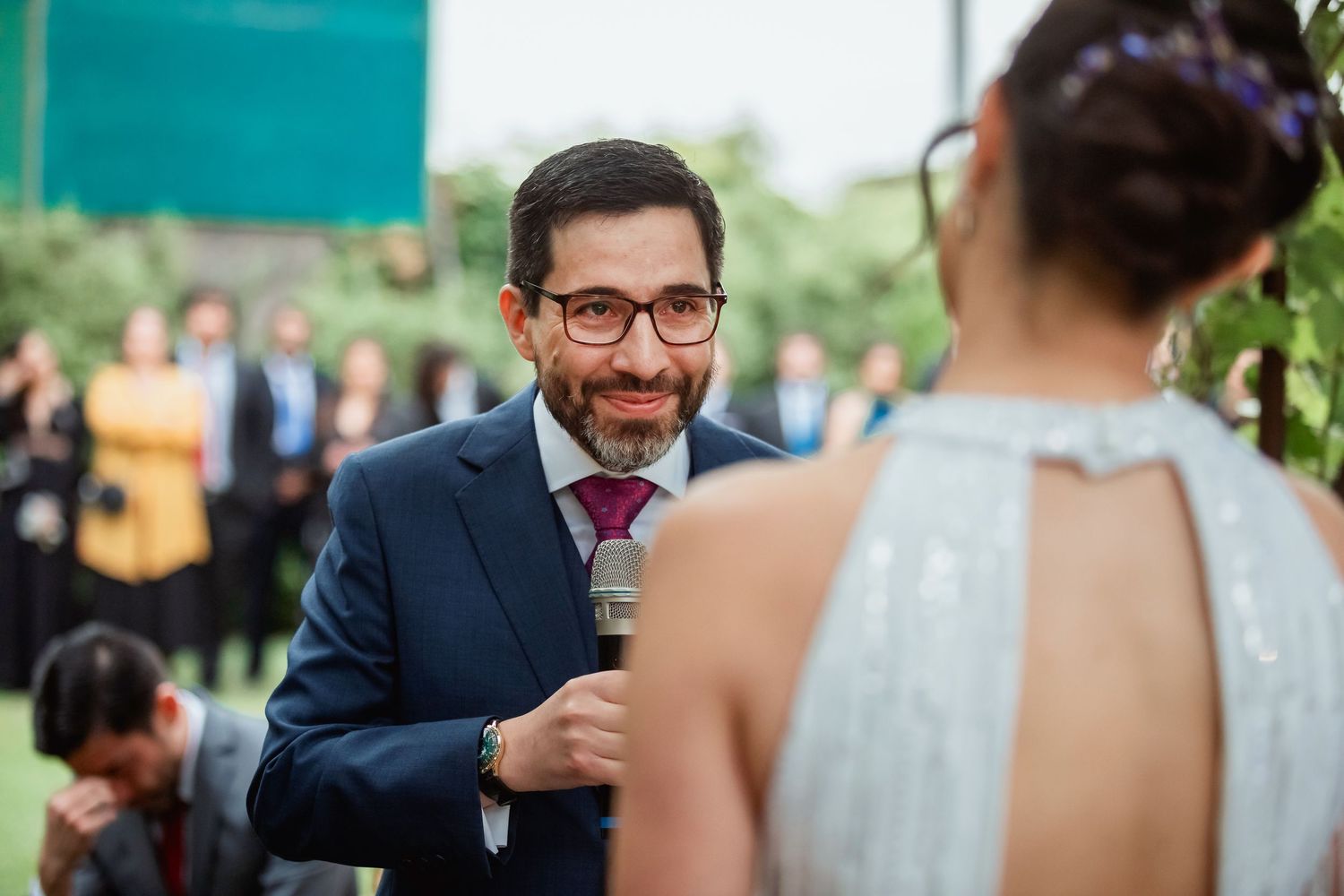 Groom in navy suit smiles warmly at bride during outdoor wedding vow exchange.