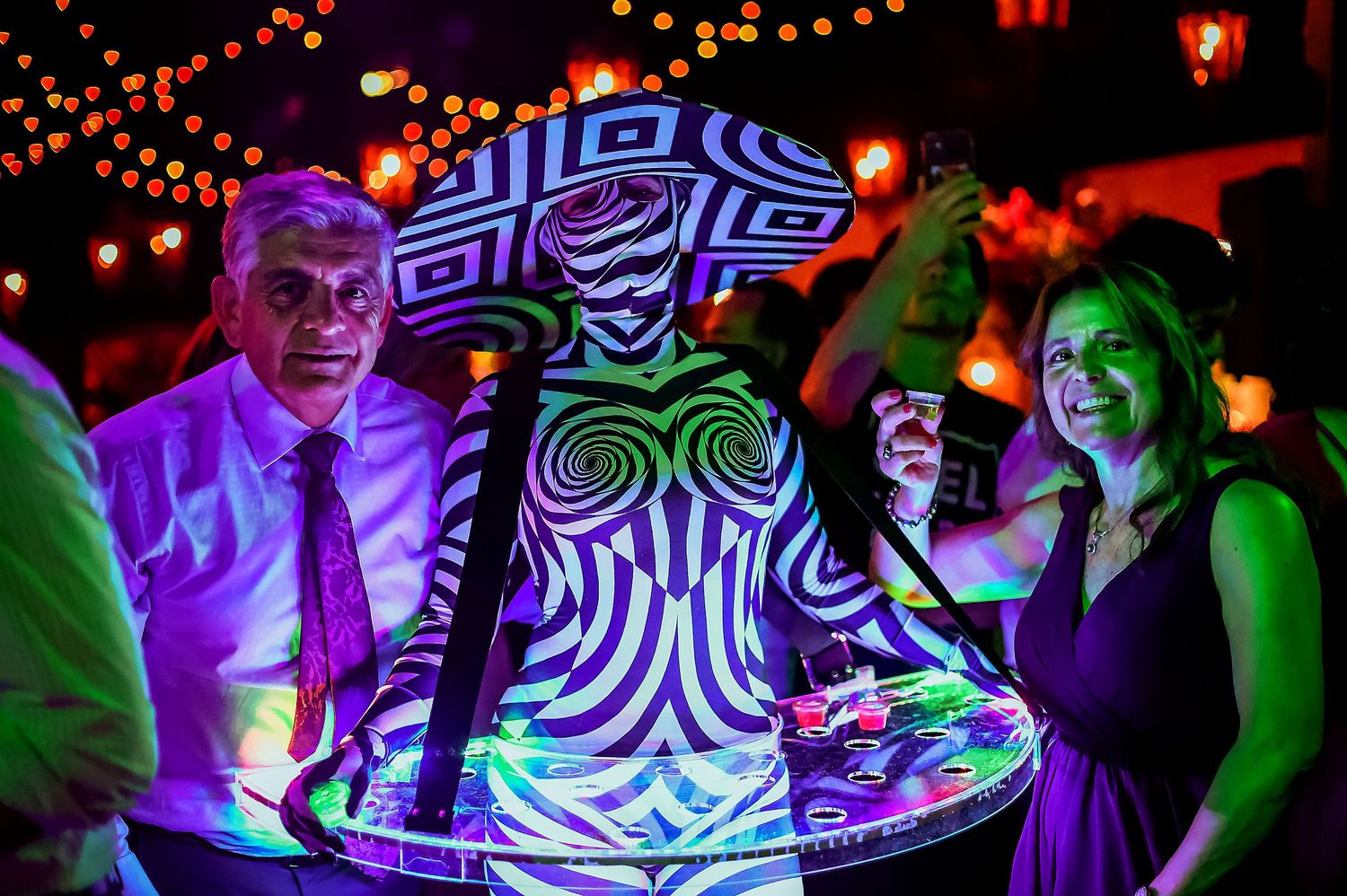 Couple poses with a zebra-print body-painted server carrying glowing drinks at a festive night event.
