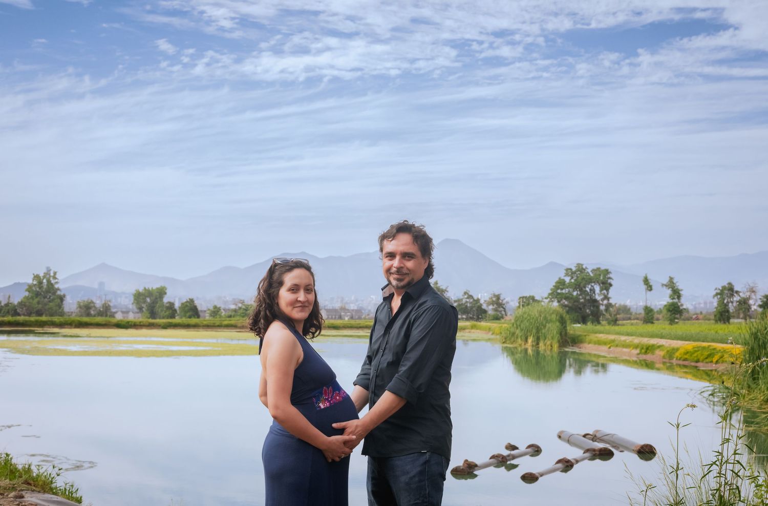 Couple standing together by calm lake with mountain backdrop under partly cloudy sky.