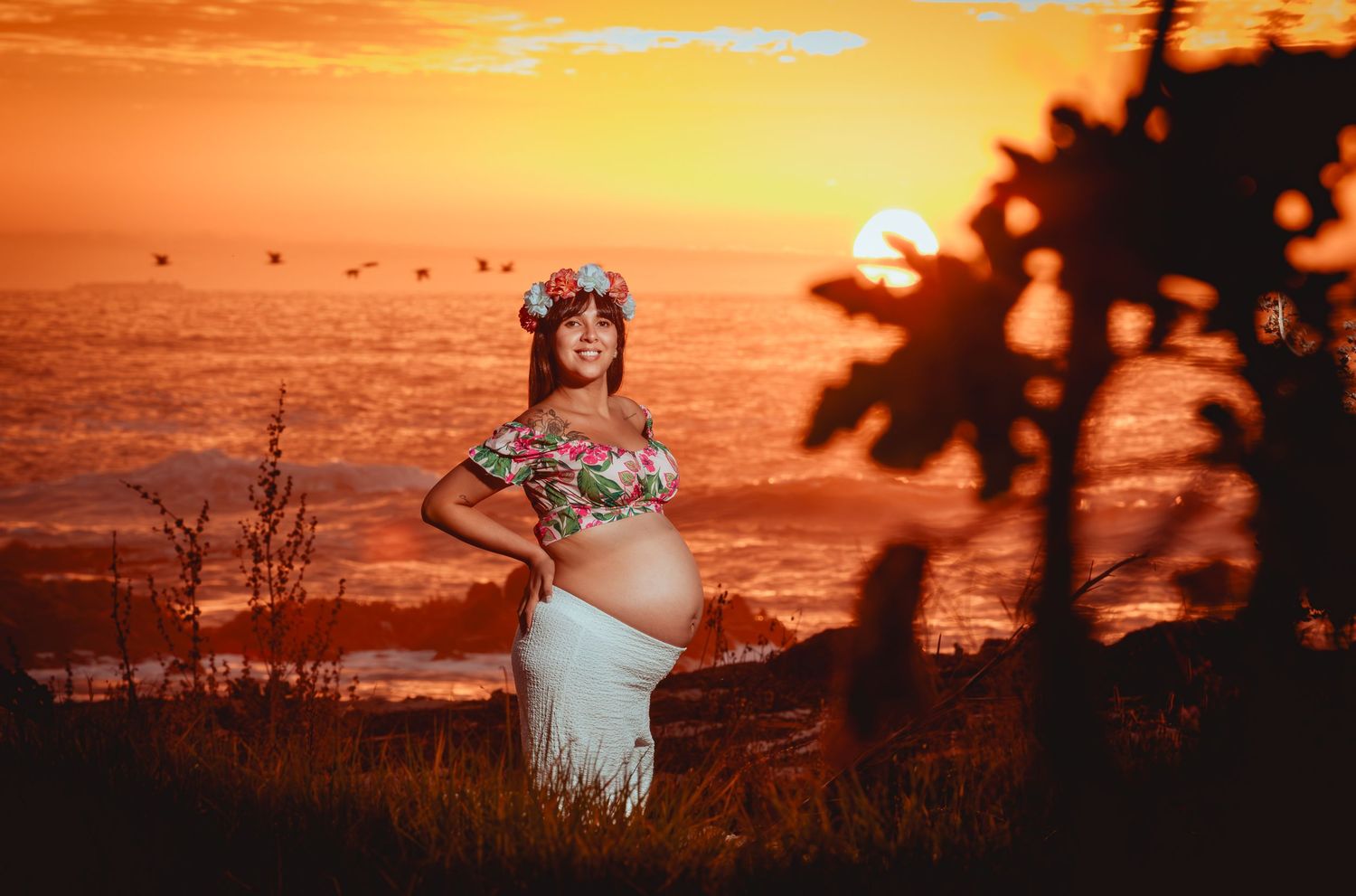 Pregnant woman in floral crop top and white skirt with flower crown poses at golden sunset over ocean.