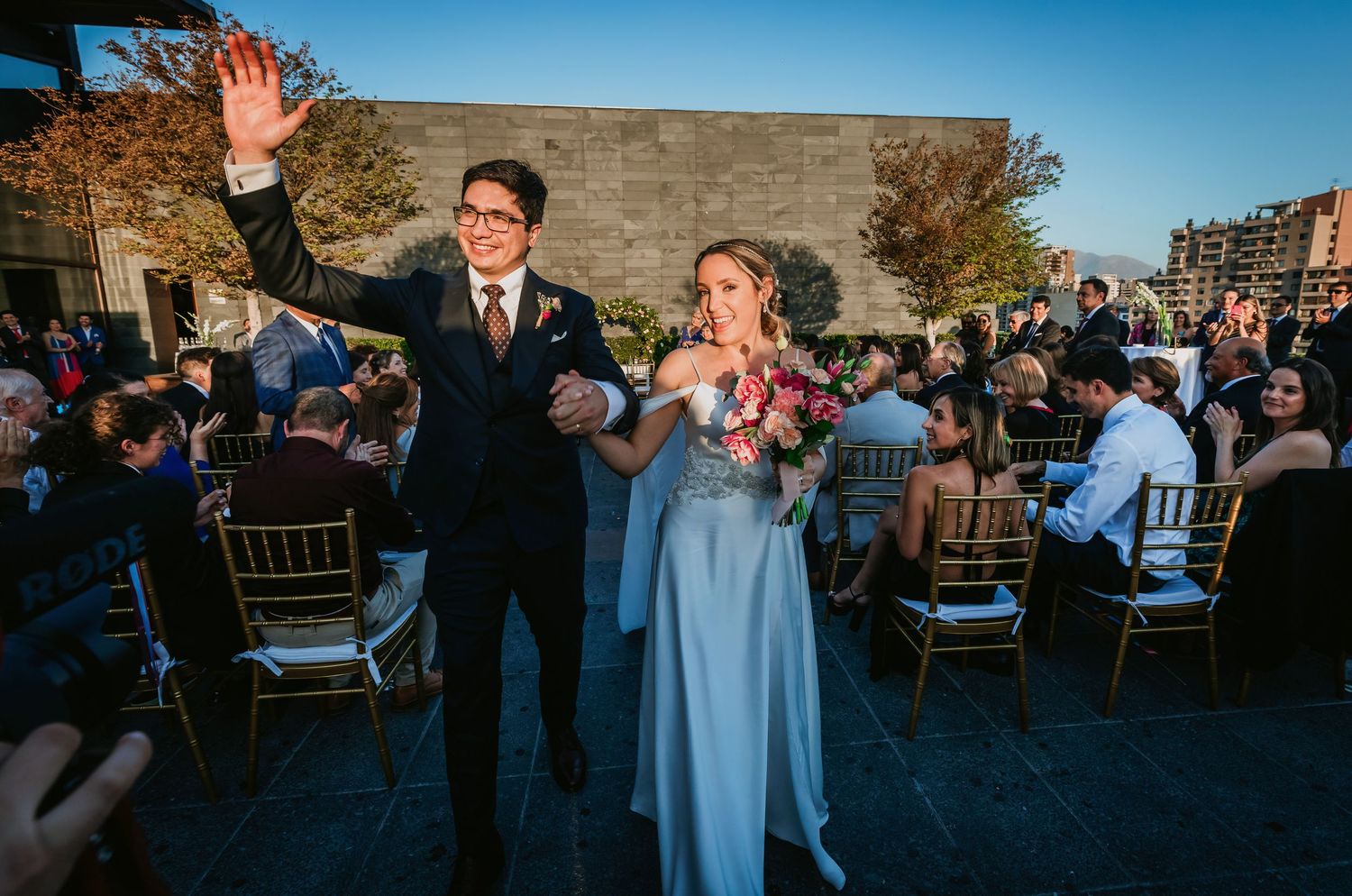 Groom waves while walking with bride in blue gown after outdoor wedding ceremony.