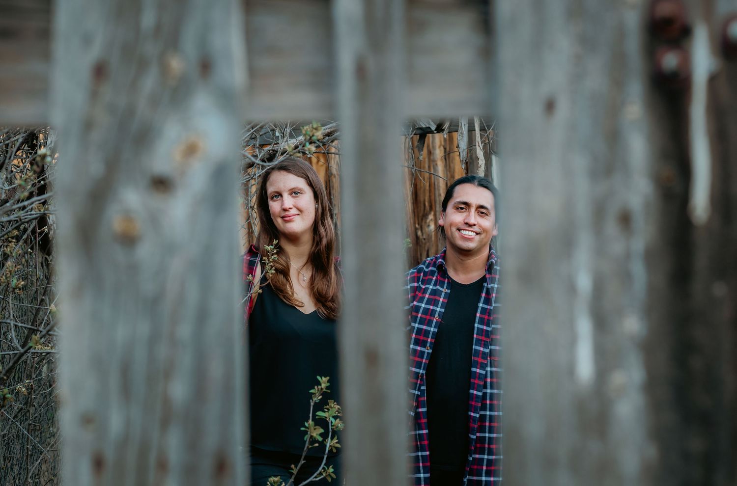 A man and woman peer through gaps in a weathered wooden fence with a rustic barn backdrop.