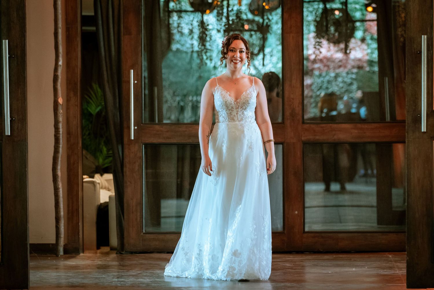 Radiant bride in flowing white lace wedding dress walks through wooden doors at wedding venue.