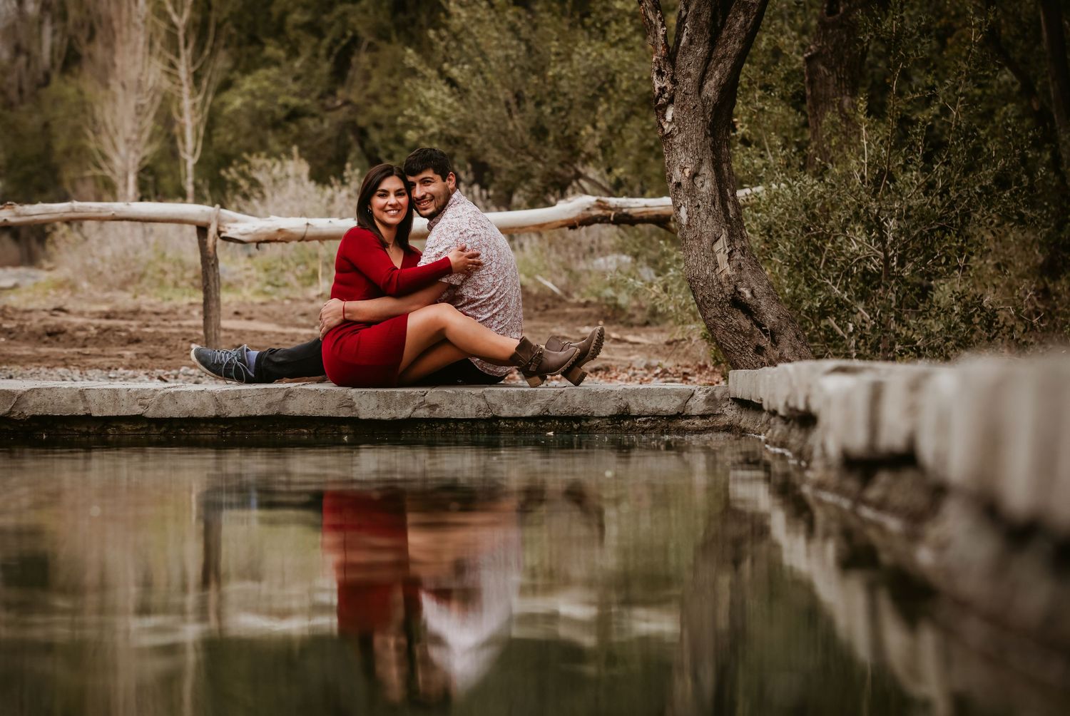 Couple sitting together on wooden dock reflected in calm water surrounded by trees in nature.
