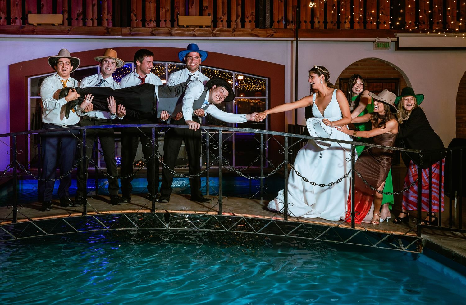 Bride and groom hold hands with wedding guests on bridge over pool at venue.
