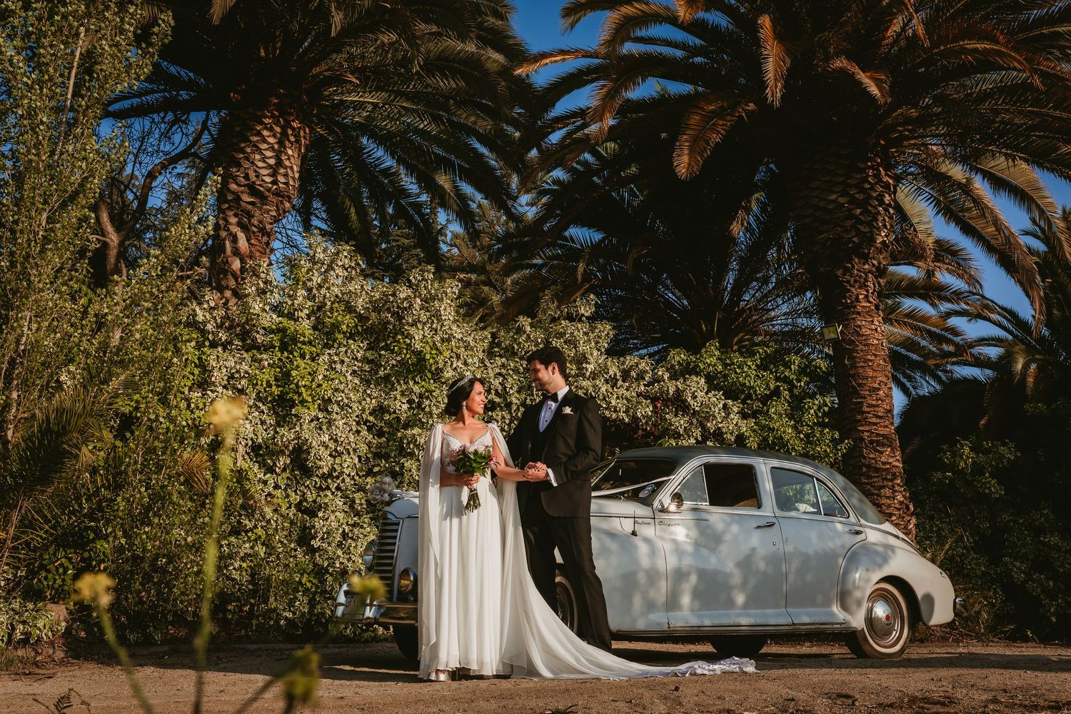 Bride and groom standing beside vintage white car surrounded by tall palm trees.