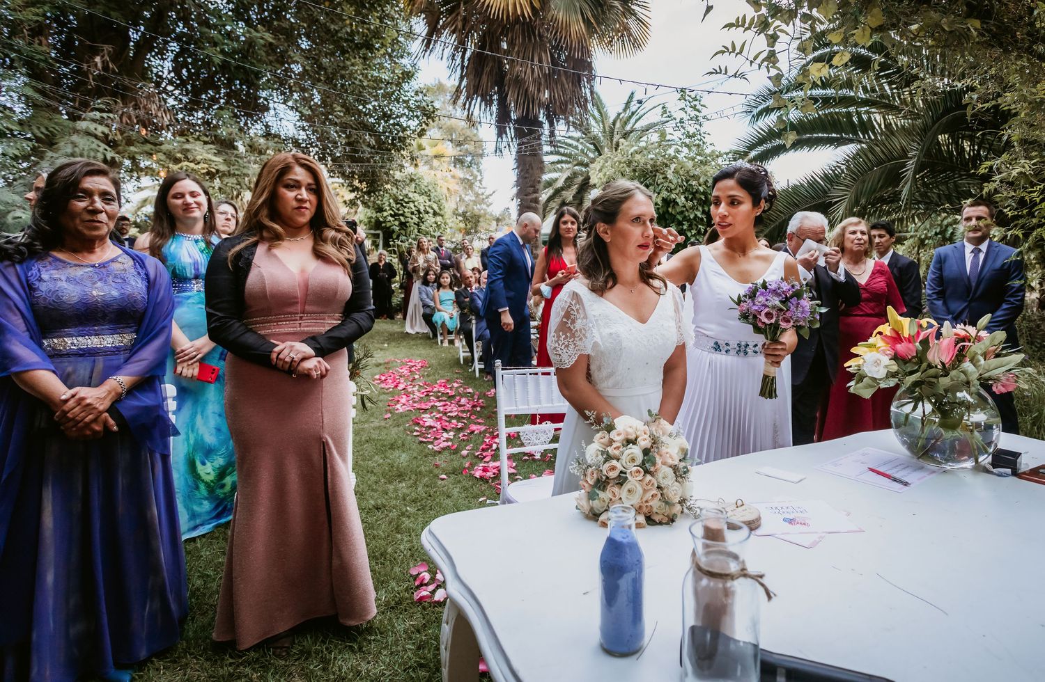 Bride stands at altar with bouquet as elegantly dressed wedding guests look on in garden ceremony.