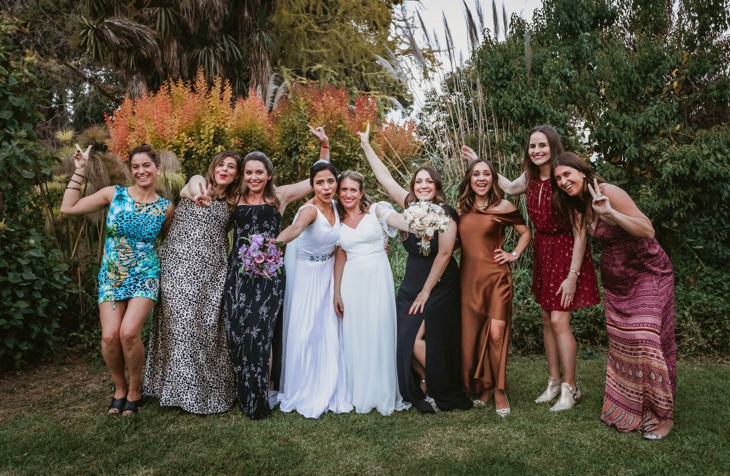 Two brides celebrate joyfully with a group of friends in a lush outdoor garden setting.