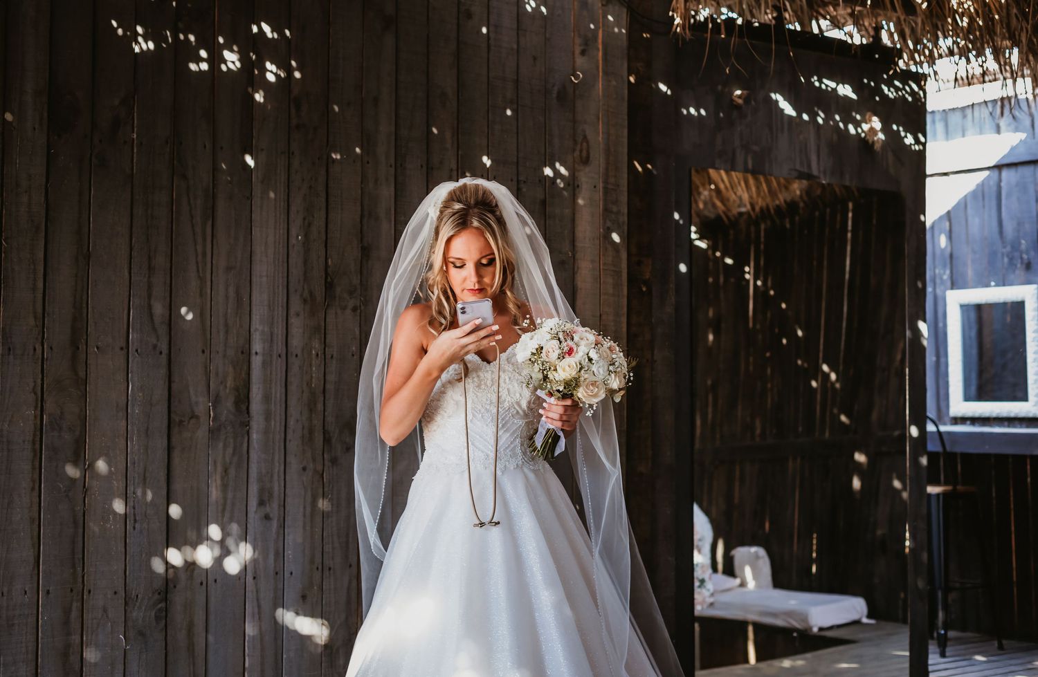 Bride in white gown with veil looking down at bouquet of roses at outdoor wedding venue.