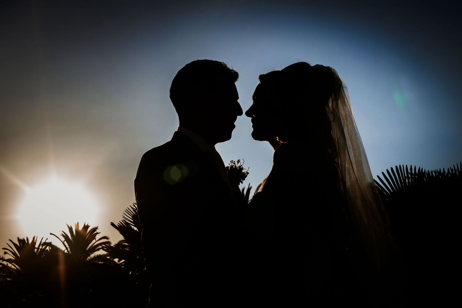 Romantic silhouette of bride and groom facing each other against glowing sunset with palm trees.