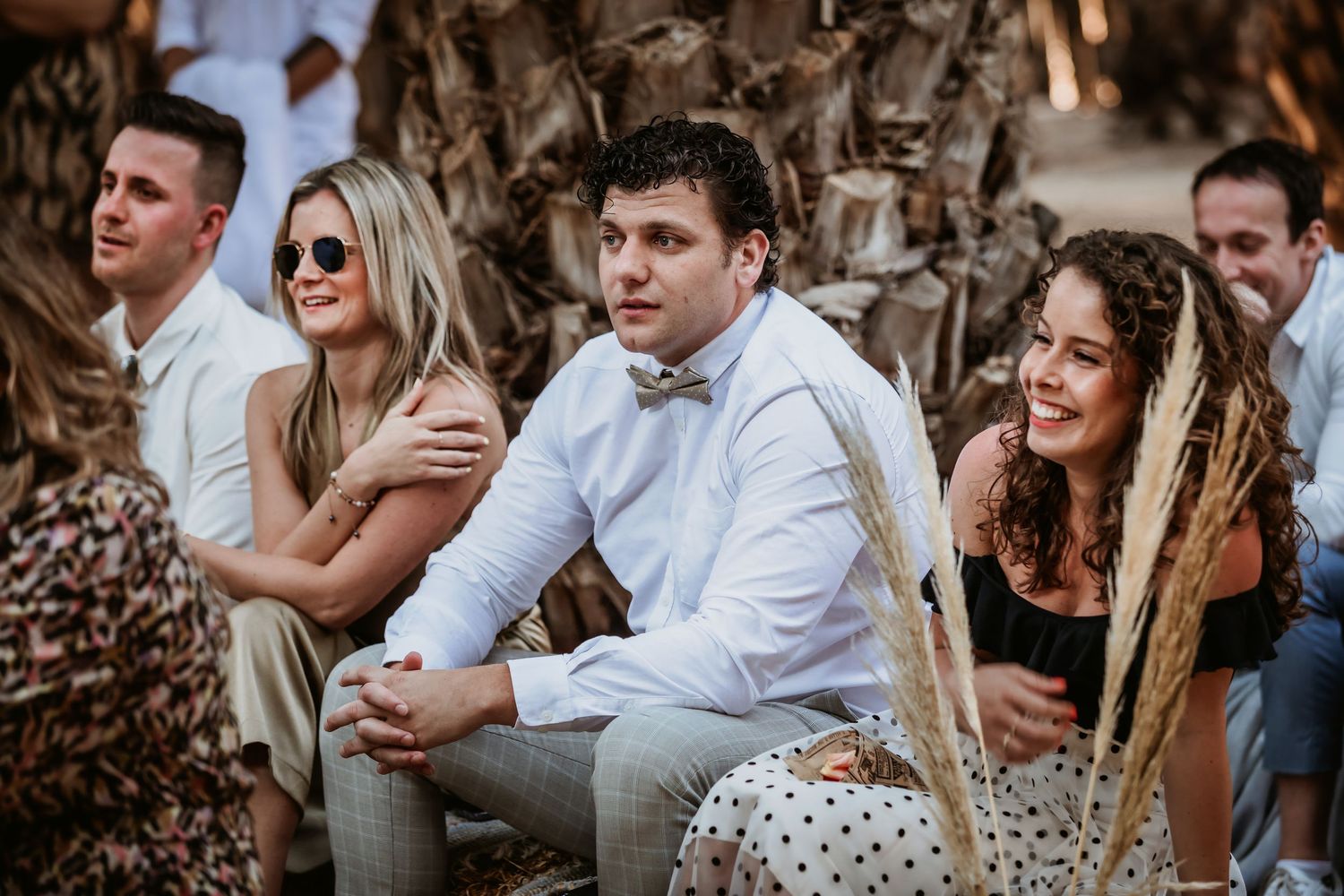 Wedding guests including man in white shirt with bow tie laugh and smile during outdoor ceremony.
