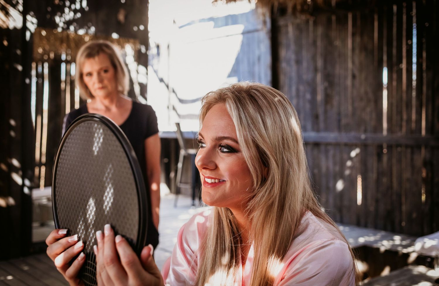 A smiling blonde bride in a pink robe examines her reflection in a round mirror held by an assistant.