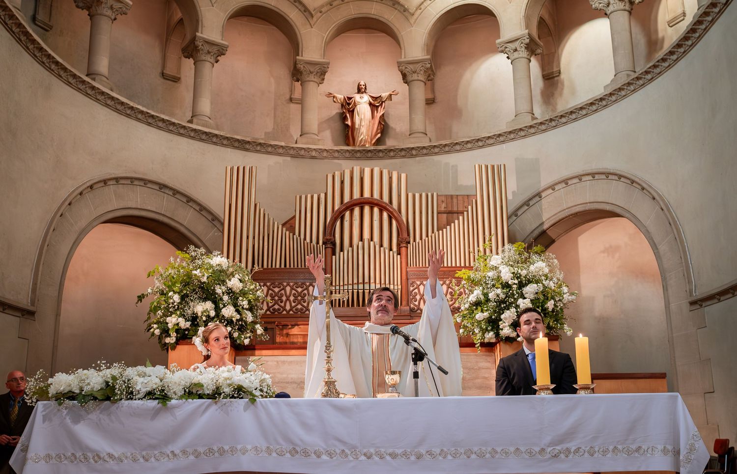 Priest raises hands at ornate church altar flanked by floral arrangements during wedding mass.