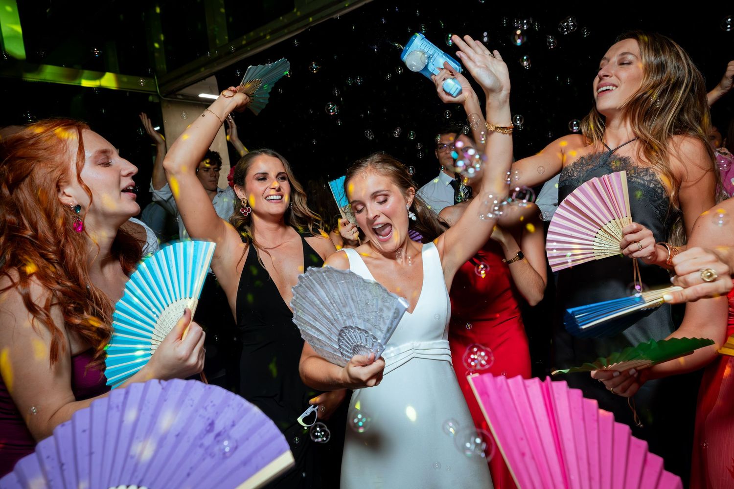 Bride and friends dancing joyfully with colorful hand fans at wedding reception.
