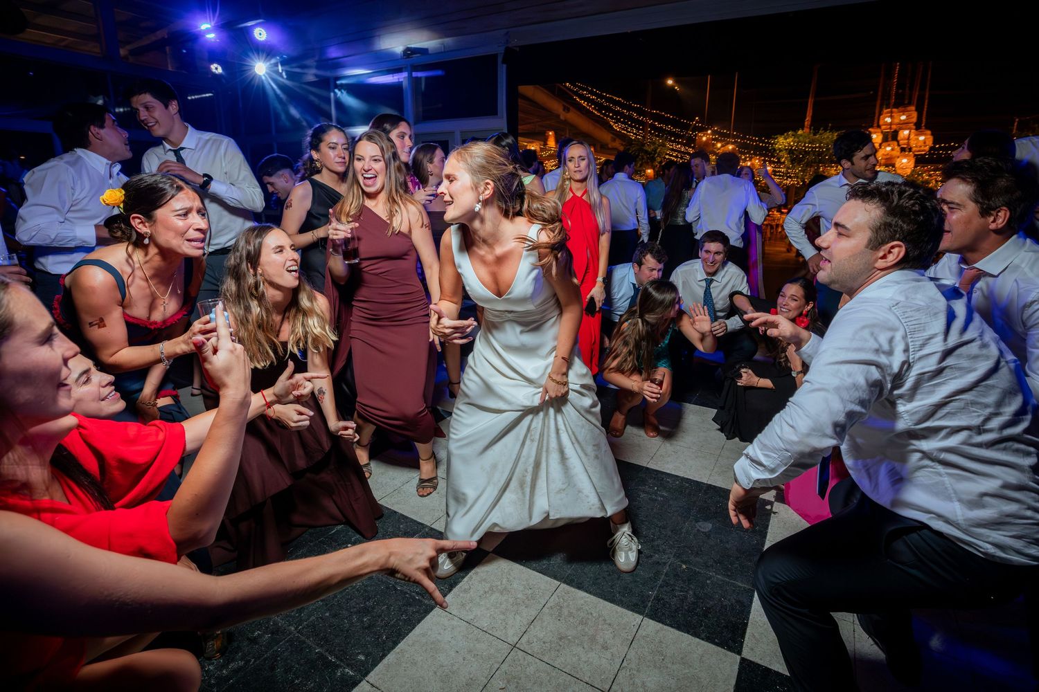 Bride and groom share energetic dance moment surrounded by cheering guests on checkered wedding reception floor.