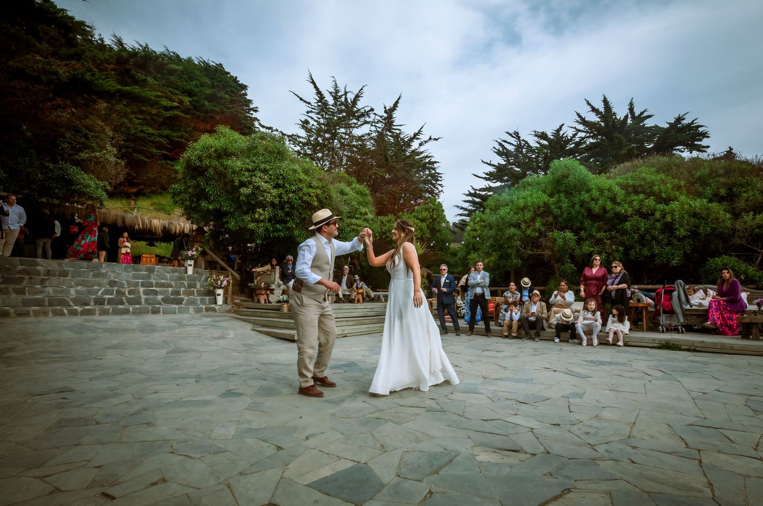 Bride and groom dancing together on a stone patio at an outdoor wedding with guests watching in the background.