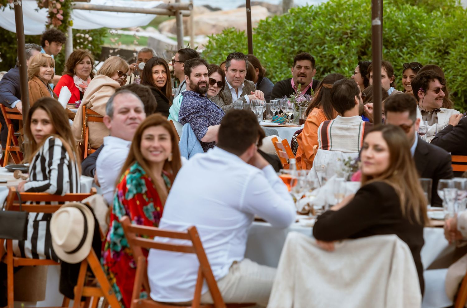 Large group of elegantly dressed guests socialize at an outdoor wedding reception with orange chairs.
