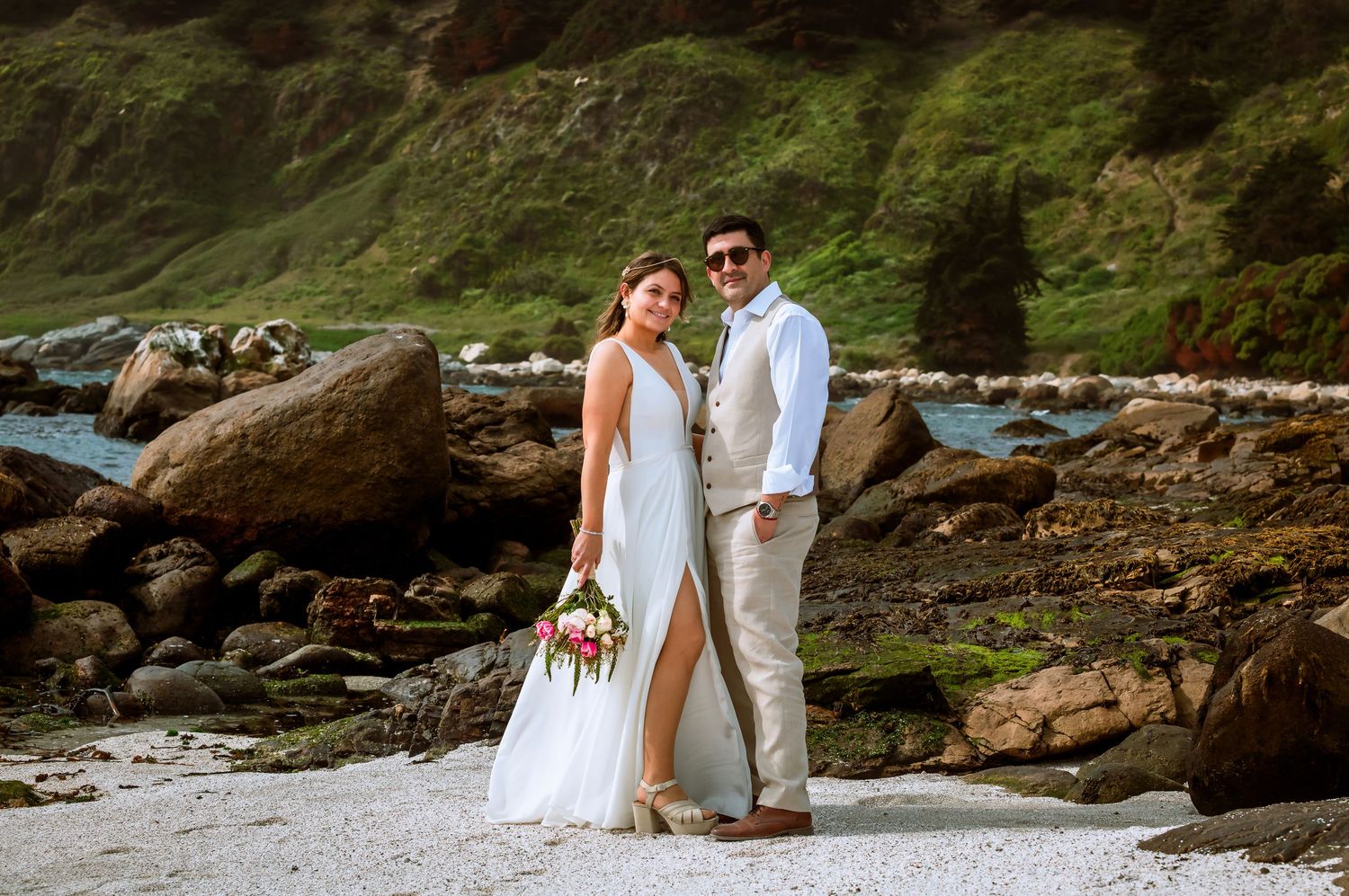 Couple in wedding attire posing on rocky beach, bride in white gown holding bouquet, groom in tan suit.