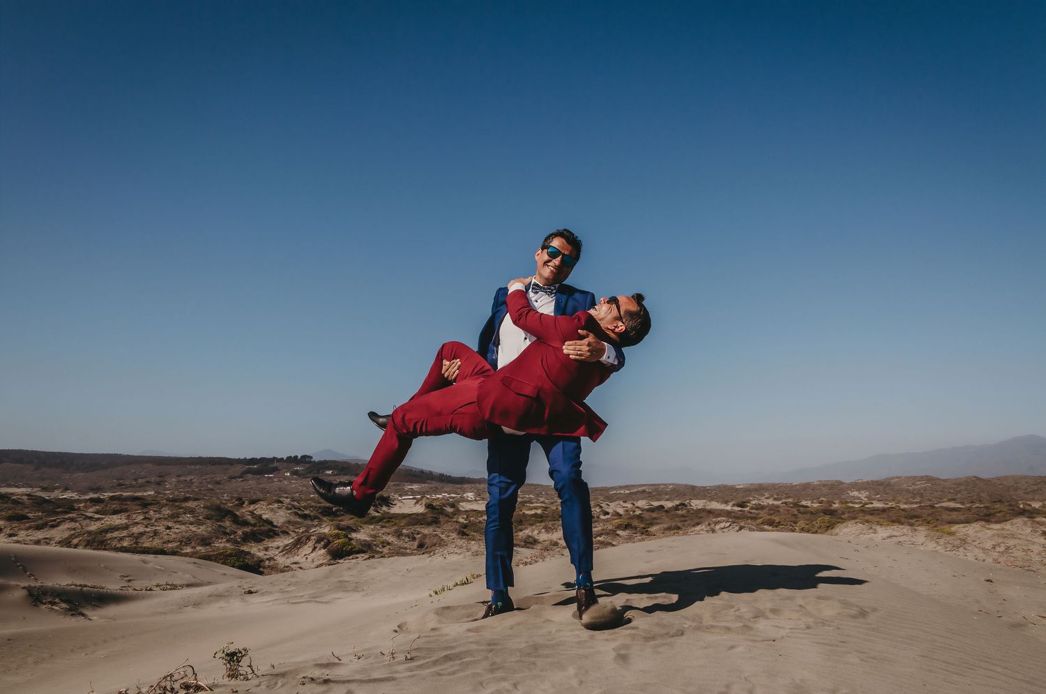 Groom in blue suit lifting partner in red suit playfully on sunny desert sand dunes.