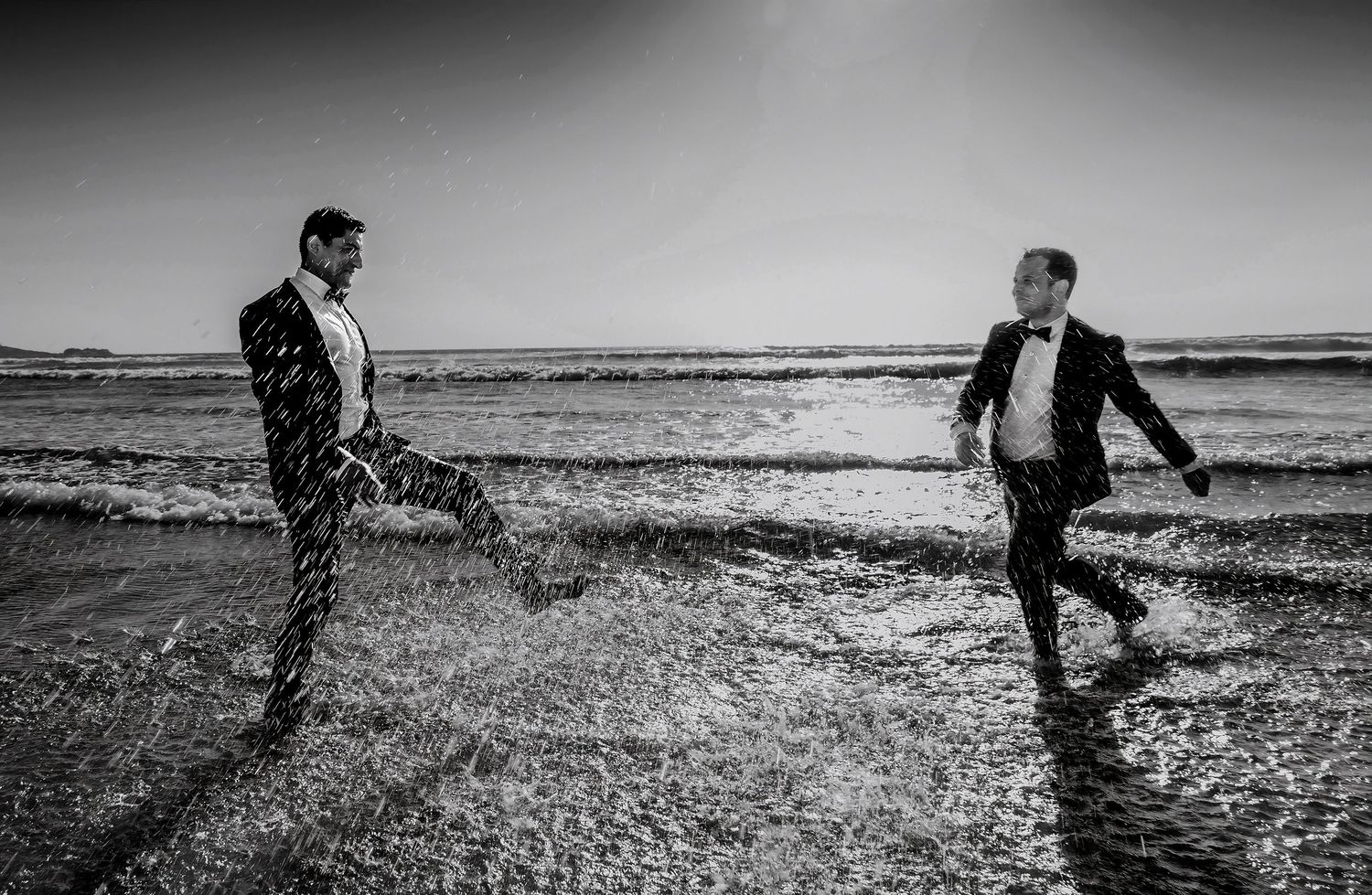 Black and white photo of two tuxedo-clad grooms splashing and running playfully along a sunny beach.
