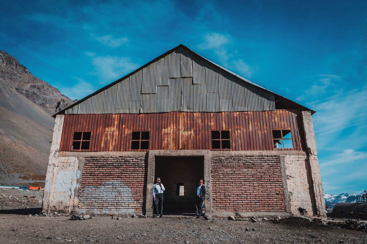 Two grooms in formal attire stand before a rustic corrugated metal barn with snow-capped mountains under blue sky.