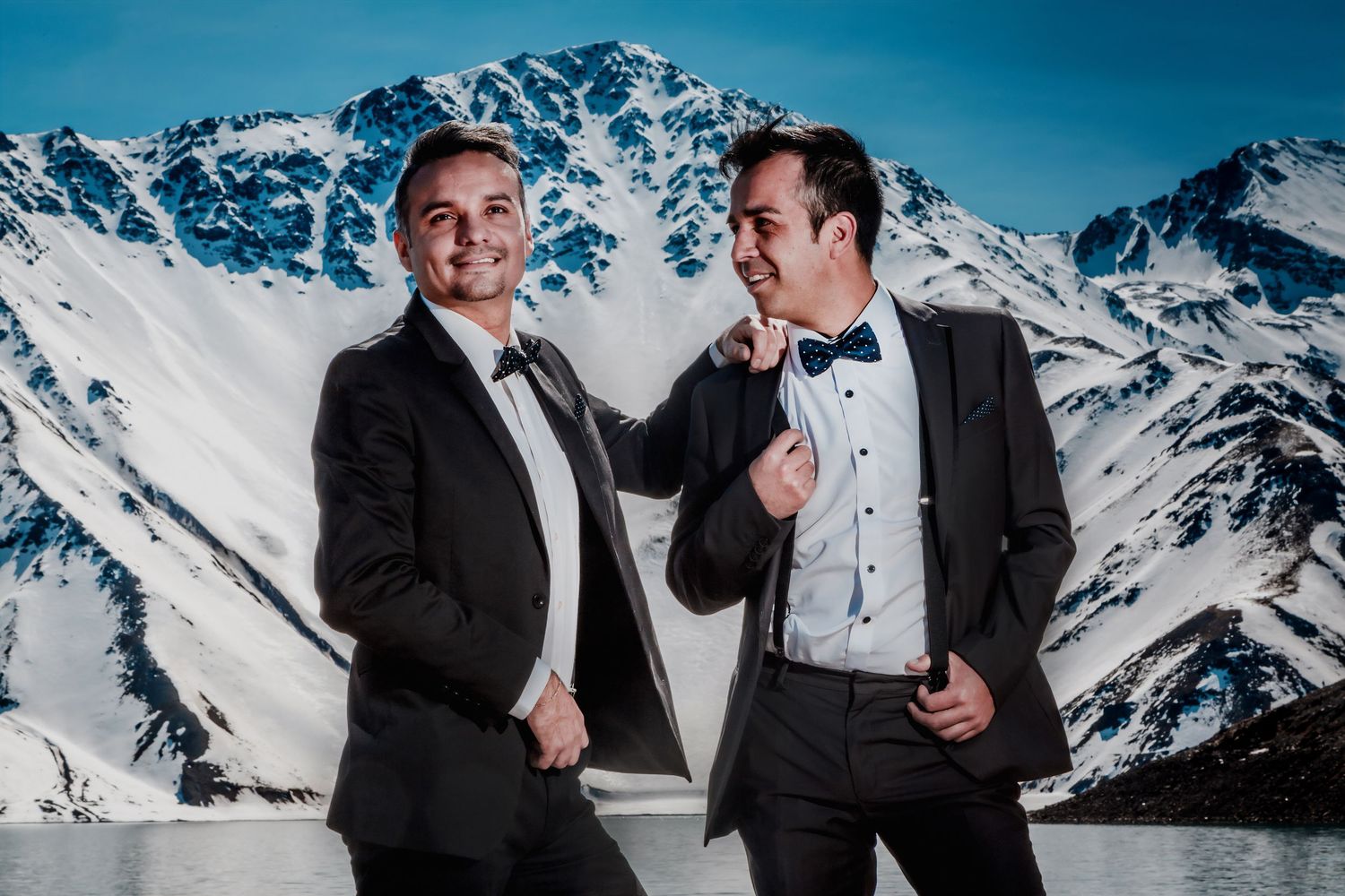 Two grooms smile and adjust each other's bow ties in front of dramatic snow-covered mountains.