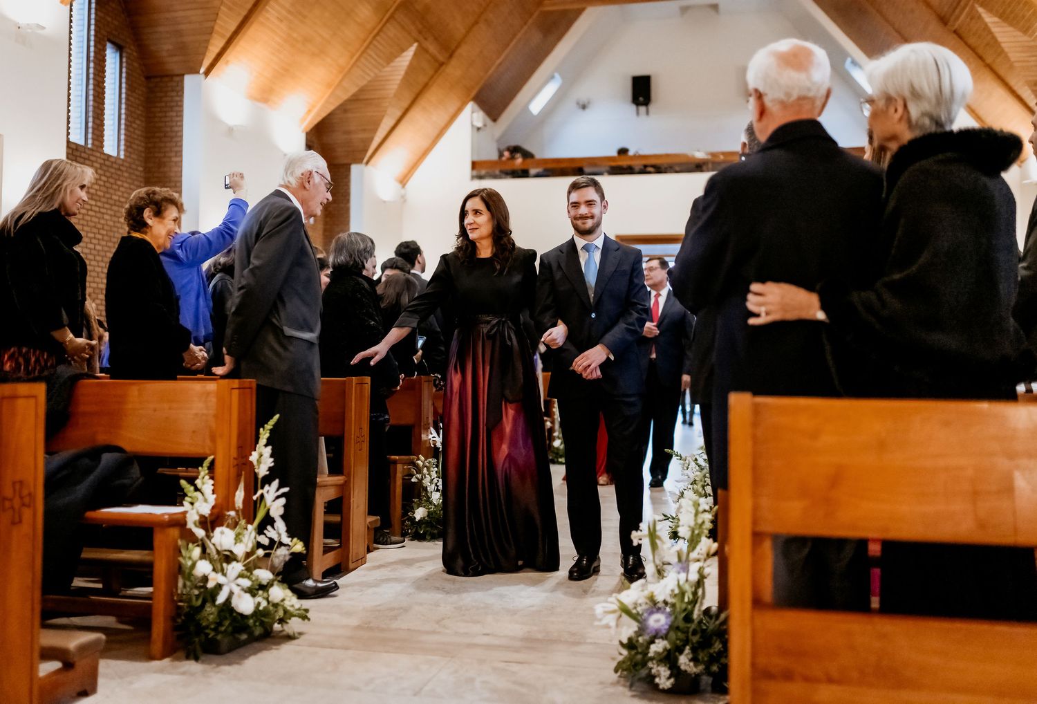 Couple walks down church aisle in formal attire during a wedding ceremony, surrounded by seated guests.