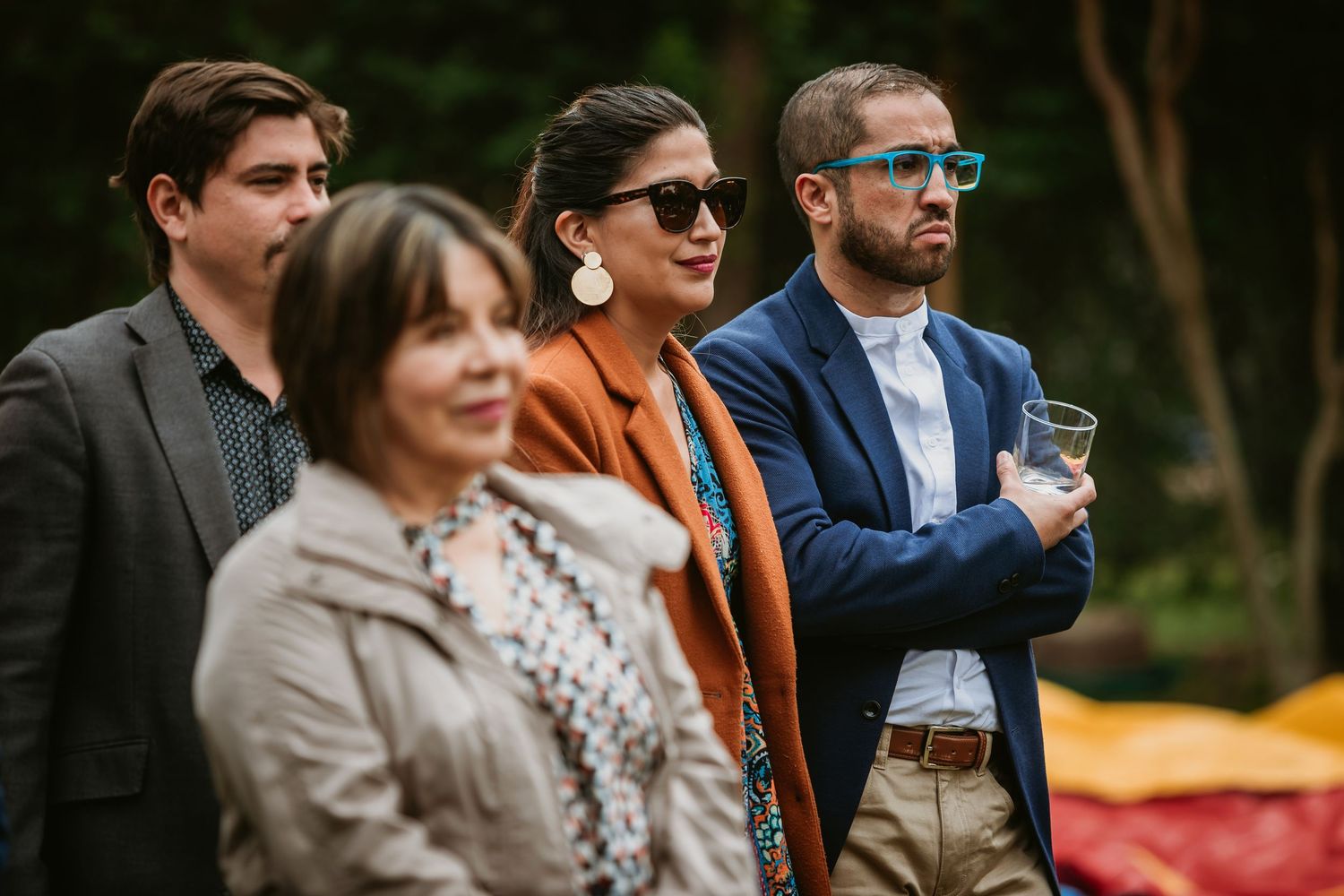 Four guests in stylish attire, some wearing sunglasses, attending an outdoor garden party or wedding reception.