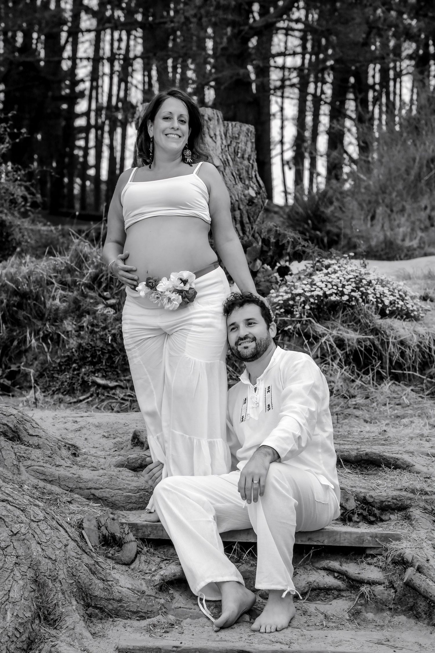 Black and white maternity photo of pregnant woman and man in white outfits posing outdoors among tree roots.