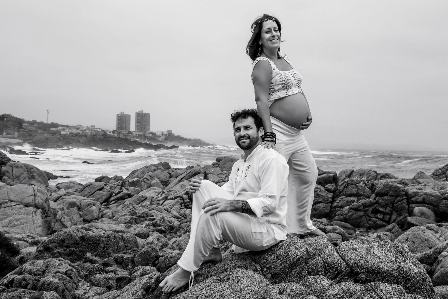 Black and white photo of a pregnant couple posing on rocky beach, man sitting while woman stands showing baby bump.