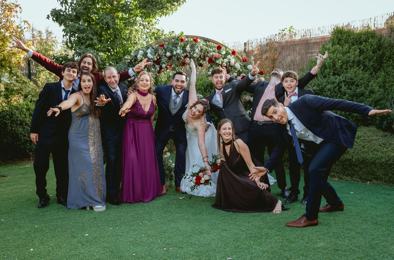 Wedding party poses joyfully with bride and groom under floral arch on green lawn outdoors.