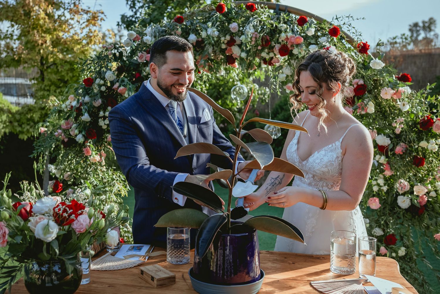 Smiling bride and groom participate in symbolic plant unity ceremony at outdoor wedding altar.