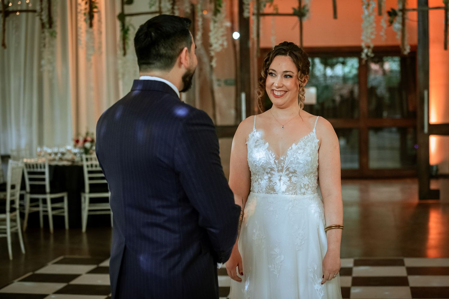 Bride in lace gown smiles joyfully at groom in navy suit during first look moment.