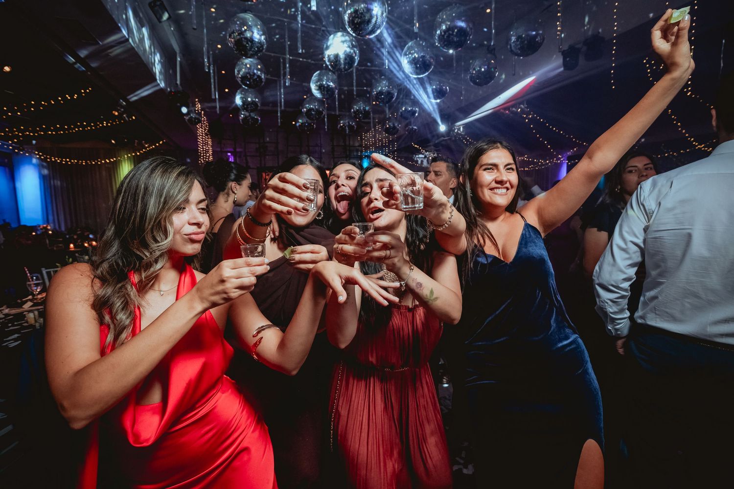Group of bridesmaids in red and blue dresses celebrate on wedding dance floor.