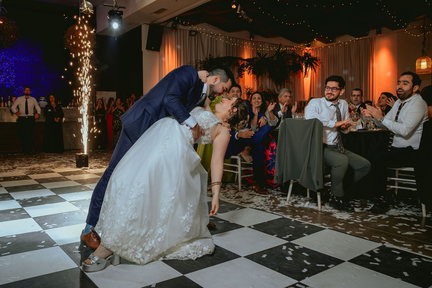 Groom dips bride dramatically on black and white checkered dance floor as wedding guests cheer and applaud.
