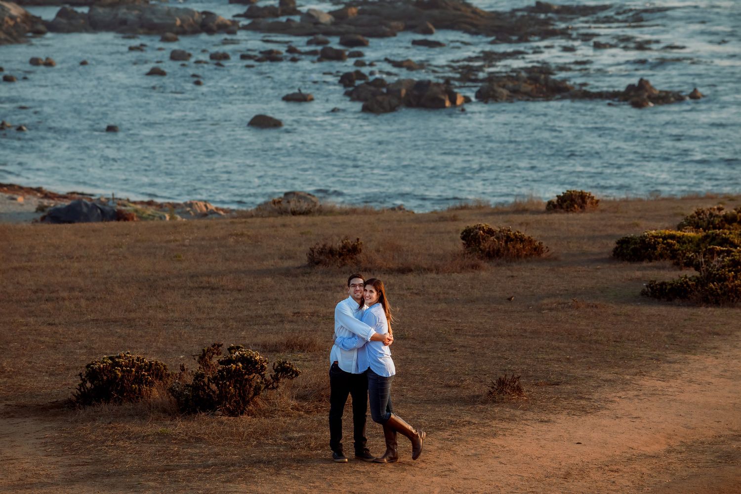 Couple embracing on a coastal hillside with rocky ocean shoreline behind them.