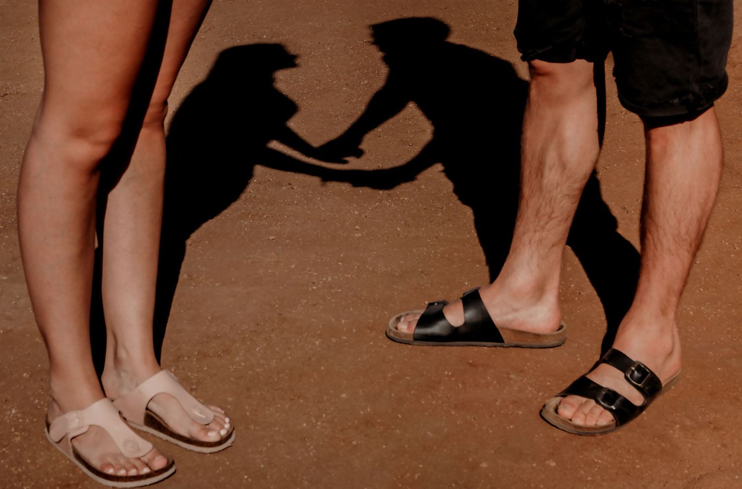 Couple's feet in sandals casting romantic shadows on sandy ground.