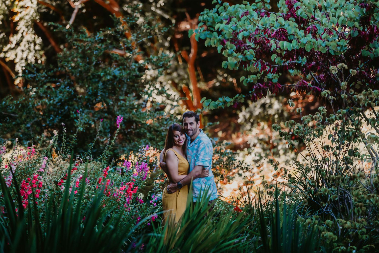 Couple embracing in a sunlit garden surrounded by colorful flowers and warm golden light.