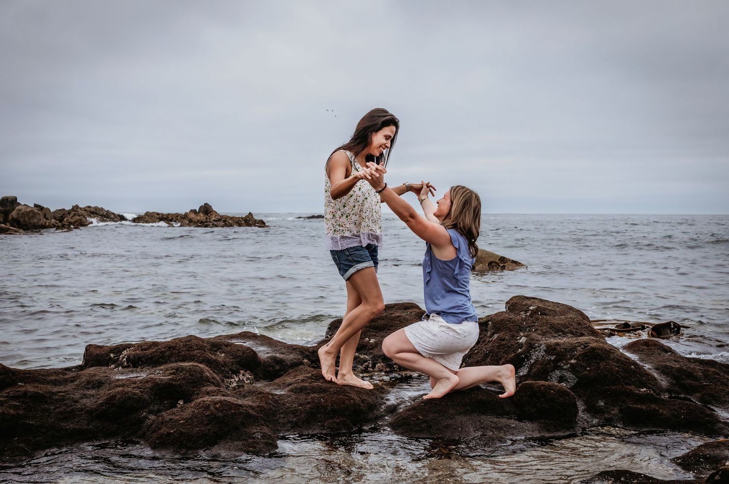 Two women playfully interact while balancing on rocky coastal boulders by the ocean shore.