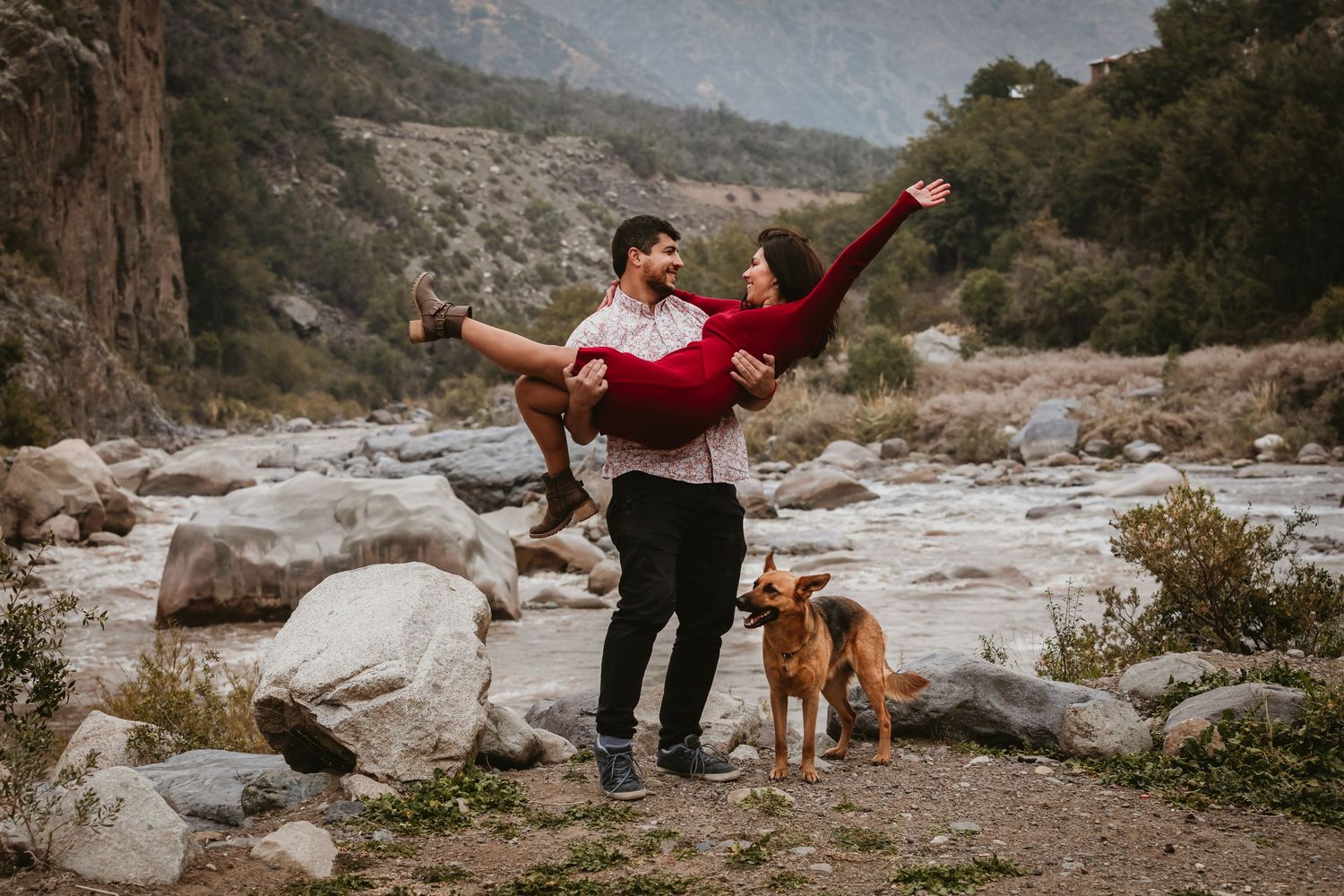 Man lifting woman in red dress outdoors by rocky river with mountains and dog watching.