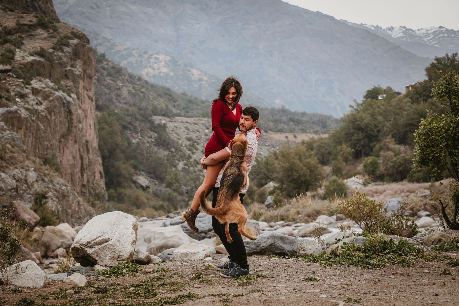 Couple playing outdoors in mountain valley, woman in red dress lifted by man with dog nearby.