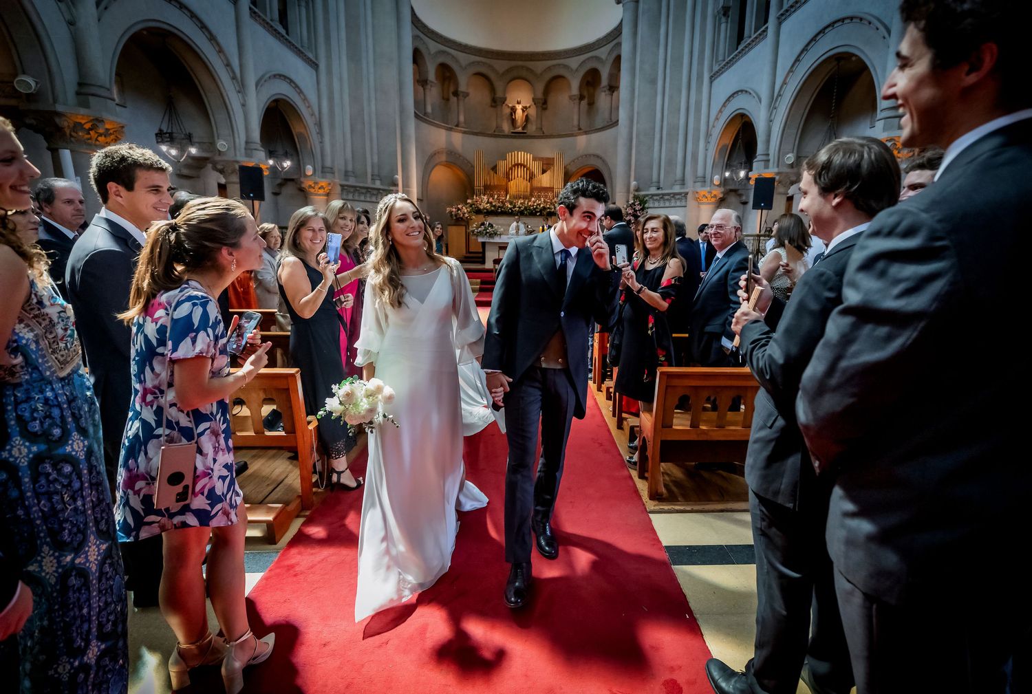 Newlyweds walk down the red carpet aisle as guests applaud in a grand church.