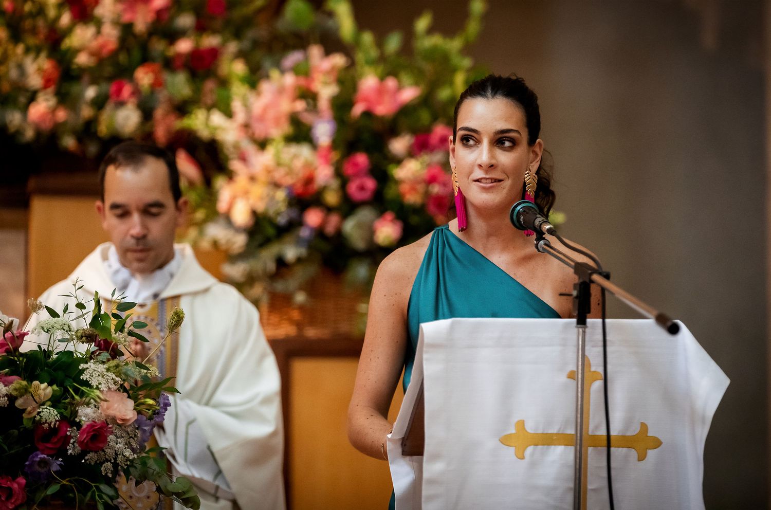 Woman in teal dress speaking at church podium during wedding ceremony with priest and flowers behind her.