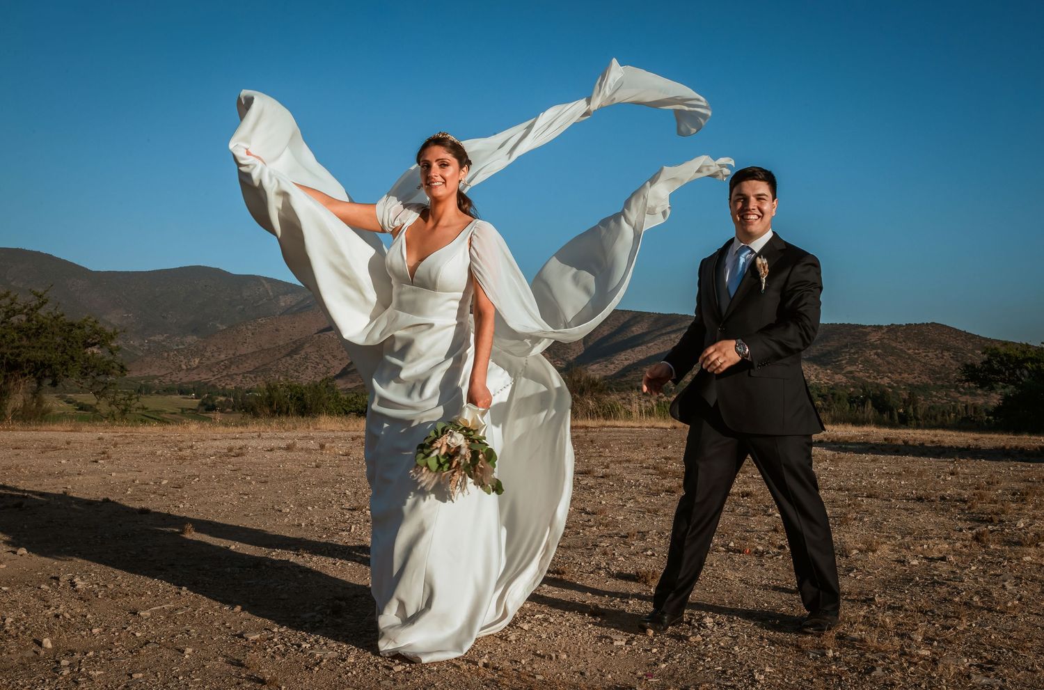 Joyful bride and groom celebrate outdoors as her long white veil soars high in the wind.
